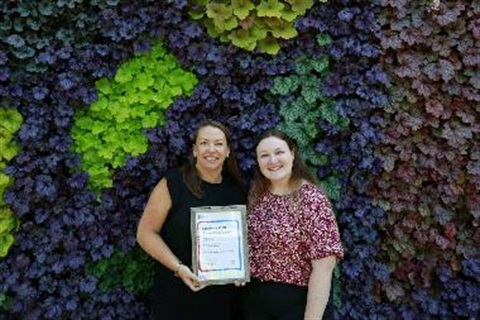 Bernadette and Eloise stand in front of a wall of foliage holding their award